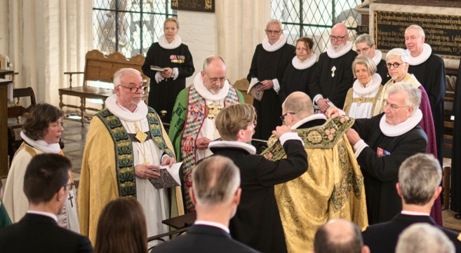 Billede af biskop Mads Davidsen, der bliver iklædt sin bispekåbe til bispevielse i Odense Domkirke. Imens ser de andre biskopper på.